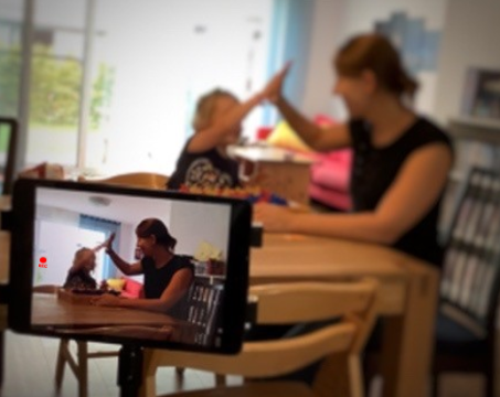Mutter und Kind am Tisch. Im Vordergrund ein Tablet mit gleicher Situation.